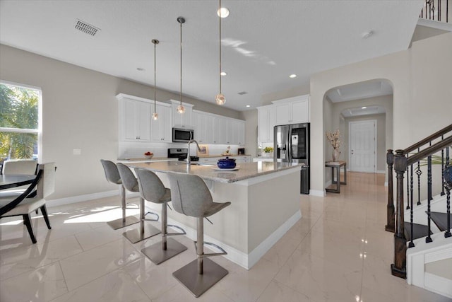 kitchen featuring white cabinetry, stainless steel appliances, a kitchen island with sink, hanging light fixtures, and light stone counters