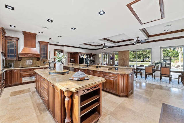 kitchen with custom exhaust hood, a center island with sink, ceiling fan, light stone countertops, and backsplash