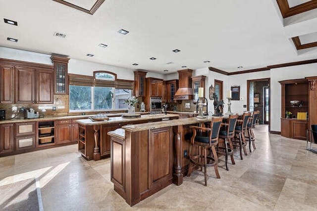 kitchen with tasteful backsplash, light stone counters, an island with sink, custom range hood, and oven