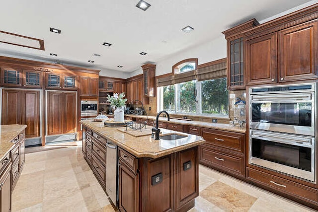 kitchen with an island with sink, light stone countertops, sink, and stainless steel double oven