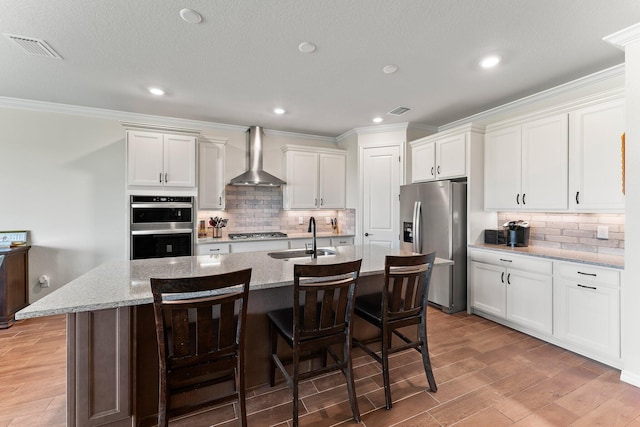 kitchen with white cabinetry, appliances with stainless steel finishes, an island with sink, wall chimney exhaust hood, and light stone counters