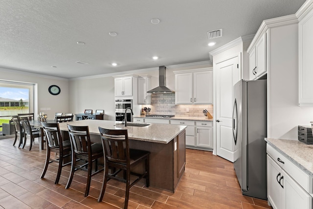 kitchen featuring a center island with sink, stainless steel appliances, wall chimney range hood, white cabinets, and sink