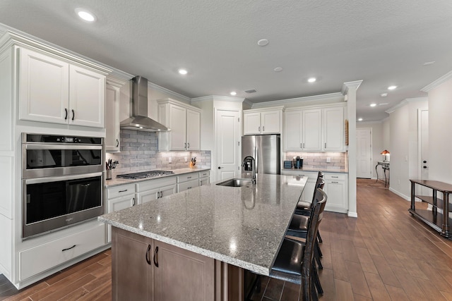 kitchen with a kitchen bar, white cabinetry, a kitchen island with sink, appliances with stainless steel finishes, and wall chimney exhaust hood