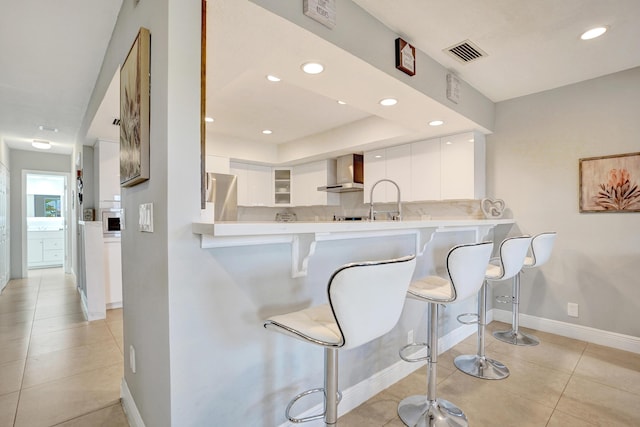 kitchen featuring white cabinetry, wall chimney range hood, a breakfast bar area, and kitchen peninsula