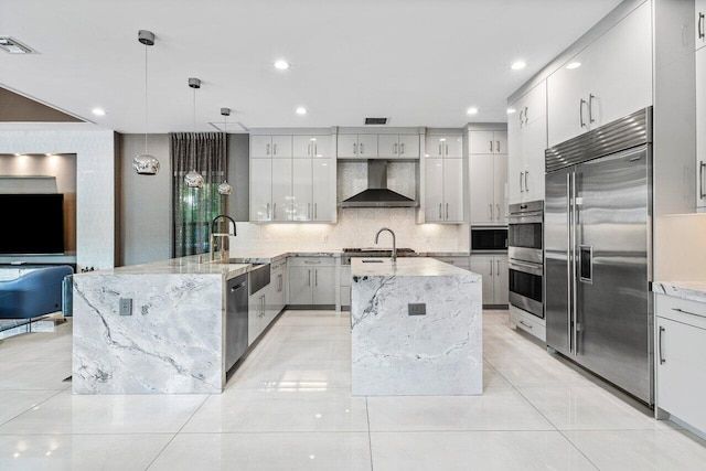 kitchen with light stone counters, decorative light fixtures, a center island with sink, stainless steel appliances, and wall chimney range hood