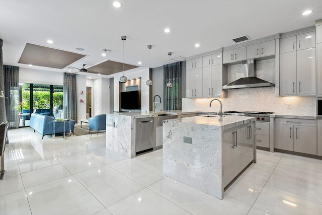 kitchen featuring wall chimney exhaust hood, pendant lighting, stainless steel appliances, light stone countertops, and a kitchen island with sink