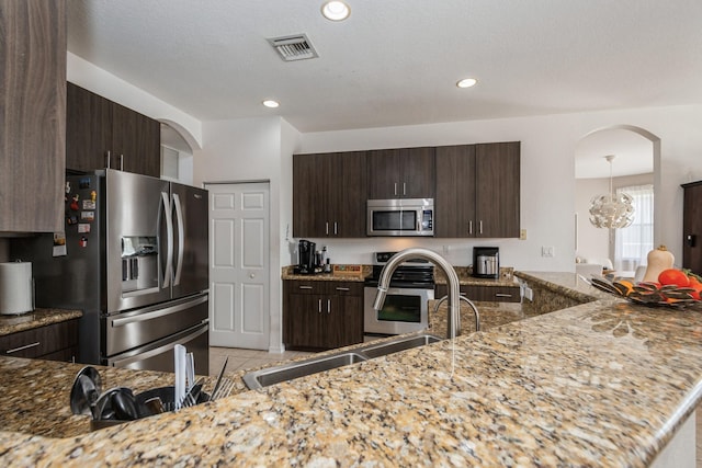 kitchen featuring appliances with stainless steel finishes, decorative light fixtures, dark brown cabinets, light stone countertops, and a chandelier