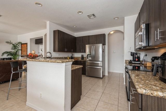 kitchen with appliances with stainless steel finishes, light stone counters, a breakfast bar, light tile patterned floors, and dark brown cabinets