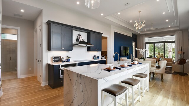 kitchen with light hardwood / wood-style floors, a center island with sink, stainless steel oven, wall chimney exhaust hood, and black electric cooktop