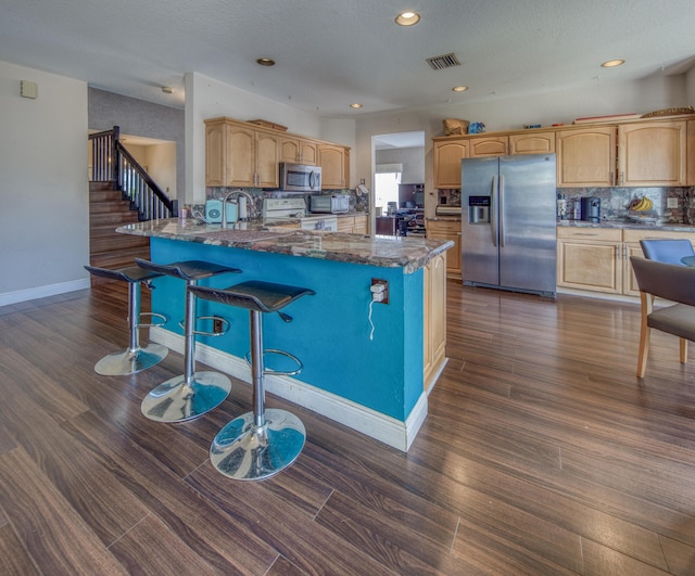 kitchen with dark wood-type flooring, a breakfast bar, appliances with stainless steel finishes, decorative backsplash, and dark stone counters