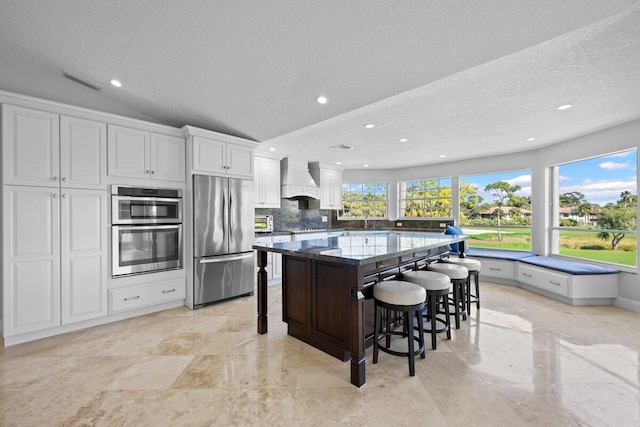 kitchen featuring dark stone countertops, premium range hood, a breakfast bar, appliances with stainless steel finishes, and white cabinets