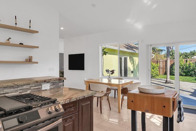 kitchen with stainless steel gas range oven, tasteful backsplash, dark brown cabinets, light tile patterned floors, and light stone countertops