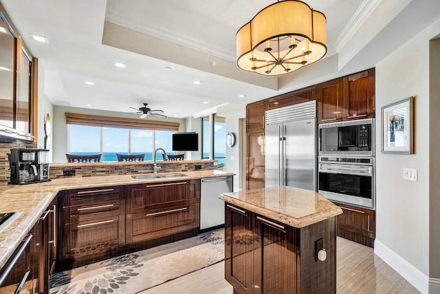 kitchen with a tray ceiling, built in appliances, crown molding, ceiling fan with notable chandelier, and sink