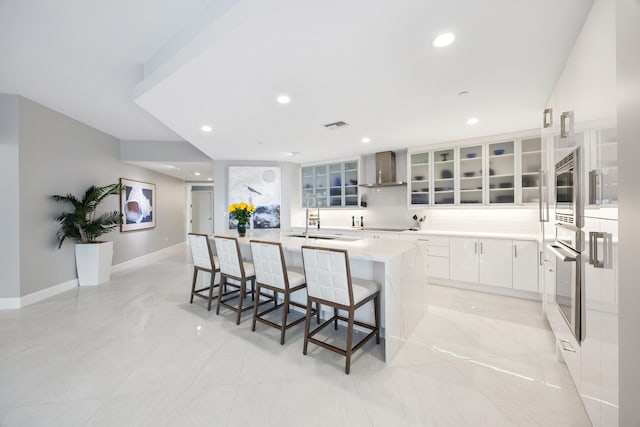 kitchen featuring a large island with sink, appliances with stainless steel finishes, wall chimney exhaust hood, a kitchen bar, and white cabinetry