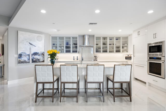 kitchen with a large island with sink, white cabinets, wall chimney range hood, built in appliances, and a kitchen breakfast bar