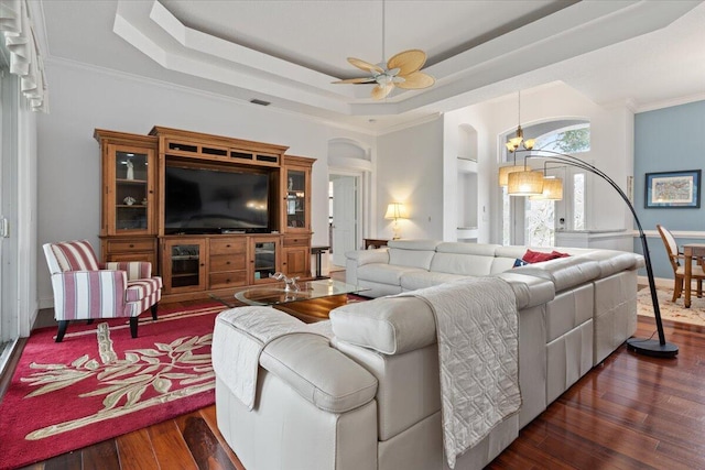 living room featuring ceiling fan with notable chandelier, dark wood-type flooring, crown molding, and a raised ceiling