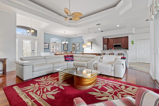 living room featuring light wood-type flooring, ornamental molding, ceiling fan with notable chandelier, and a raised ceiling