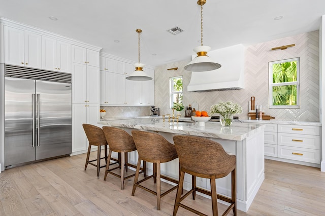 kitchen featuring built in fridge, an island with sink, pendant lighting, and white cabinetry