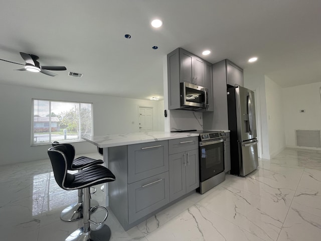 kitchen with appliances with stainless steel finishes, ceiling fan, gray cabinets, light stone counters, and a breakfast bar