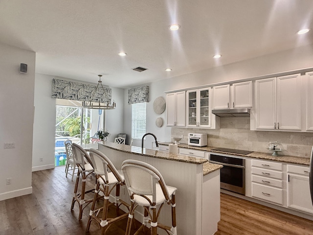 kitchen featuring white cabinetry, a center island with sink, stainless steel oven, light stone countertops, and pendant lighting