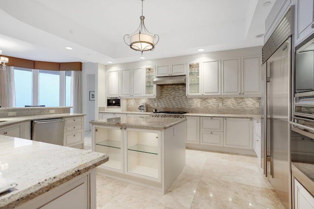 kitchen with appliances with stainless steel finishes, a raised ceiling, decorative light fixtures, light stone countertops, and a center island