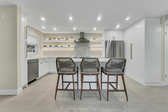 kitchen featuring a kitchen island, white cabinetry, wall chimney range hood, stainless steel appliances, and a breakfast bar area