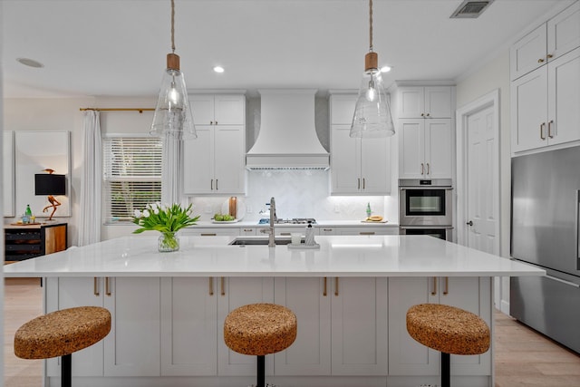 kitchen featuring premium range hood, an island with sink, a kitchen breakfast bar, decorative backsplash, and stainless steel appliances