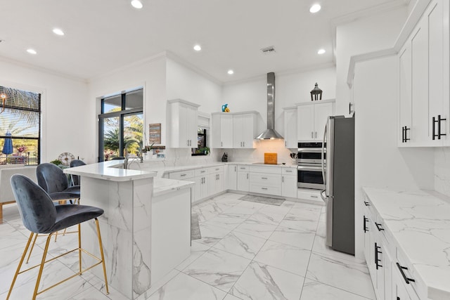 kitchen with appliances with stainless steel finishes, white cabinetry, a breakfast bar area, kitchen peninsula, and wall chimney range hood