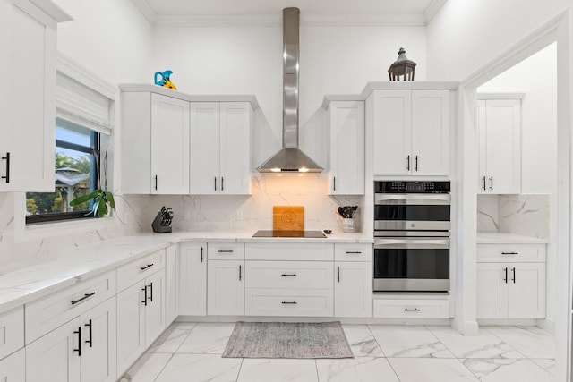 kitchen with white cabinetry, crown molding, light stone counters, double oven, and wall chimney range hood