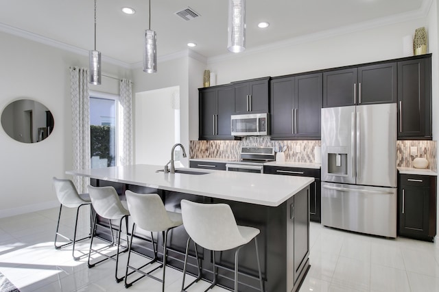 kitchen with sink, crown molding, appliances with stainless steel finishes, hanging light fixtures, and an island with sink