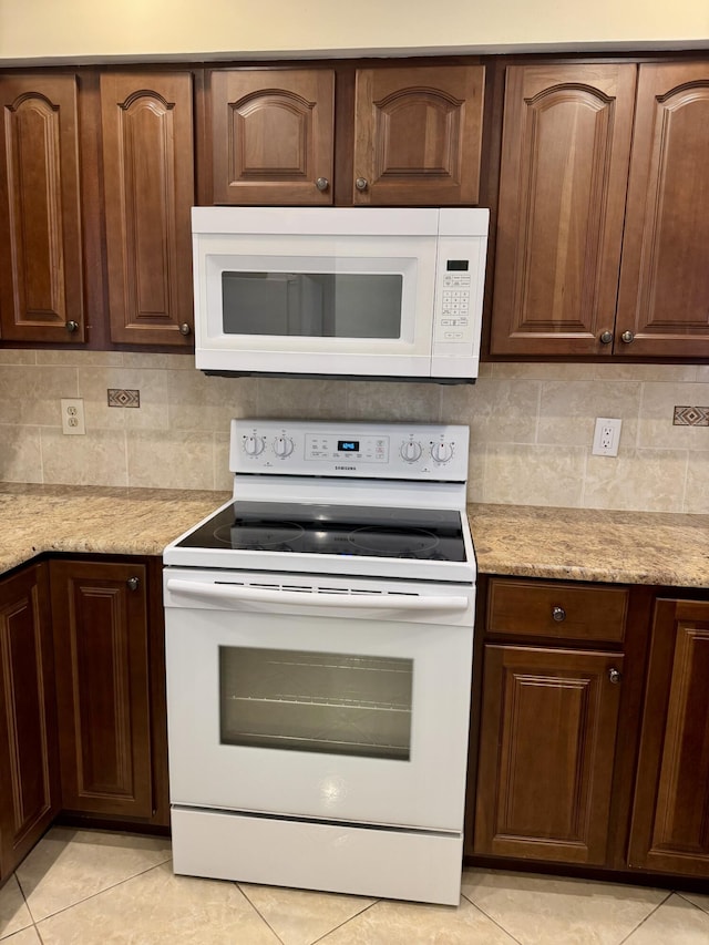 kitchen featuring decorative backsplash, light tile patterned floors, light stone counters, dark brown cabinets, and white appliances