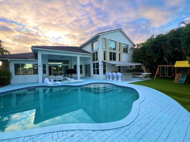 pool at dusk with a playground, ceiling fan, a lawn, and a patio