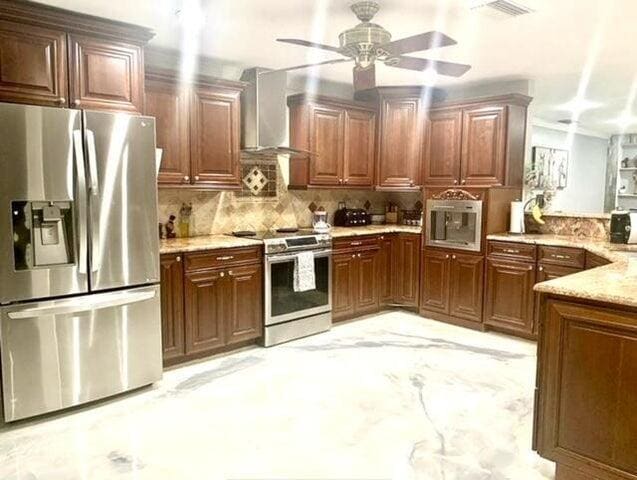 kitchen featuring appliances with stainless steel finishes, wall chimney exhaust hood, tasteful backsplash, ceiling fan, and light stone counters