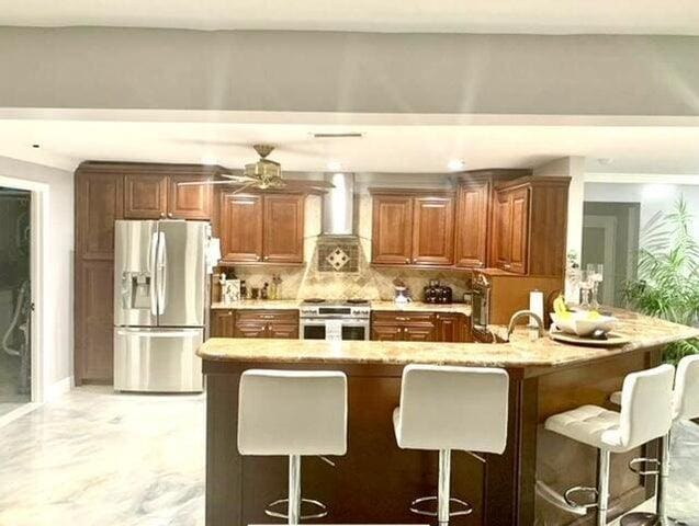 kitchen featuring kitchen peninsula, decorative backsplash, a kitchen breakfast bar, stainless steel appliances, and wall chimney exhaust hood