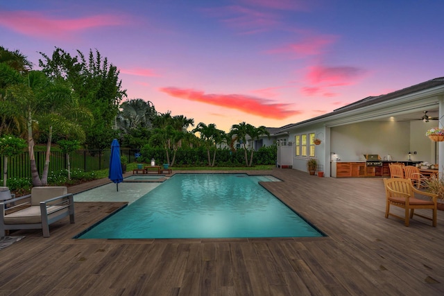 pool at dusk featuring a wooden deck, a grill, exterior kitchen, and ceiling fan