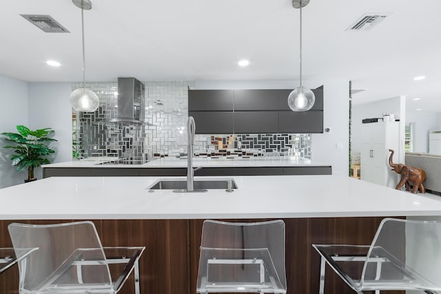 kitchen featuring backsplash, wall chimney range hood, sink, hanging light fixtures, and a kitchen island with sink