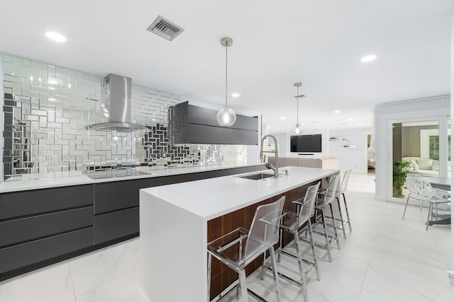 kitchen featuring tasteful backsplash, a kitchen island with sink, pendant lighting, wall chimney exhaust hood, and sink