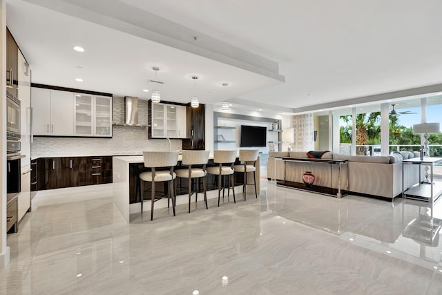 kitchen with dark brown cabinetry, a center island, hanging light fixtures, wall chimney range hood, and backsplash