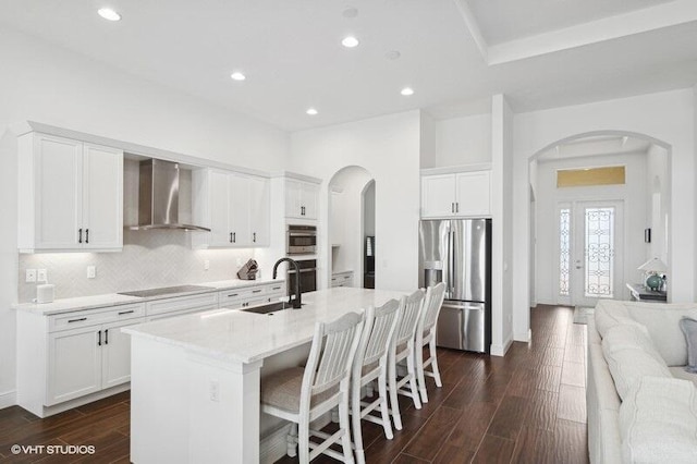 kitchen with stainless steel fridge with ice dispenser, a kitchen island with sink, wall chimney range hood, white cabinets, and sink