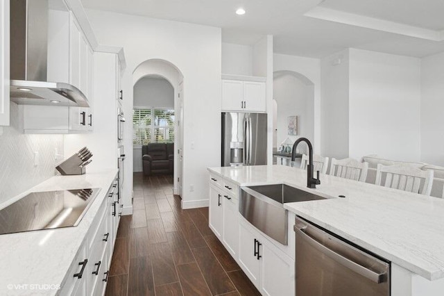 kitchen with wall chimney range hood, sink, an island with sink, stainless steel appliances, and white cabinets