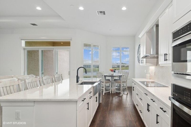 kitchen featuring a kitchen island with sink, light stone counters, black electric stovetop, and wall chimney exhaust hood