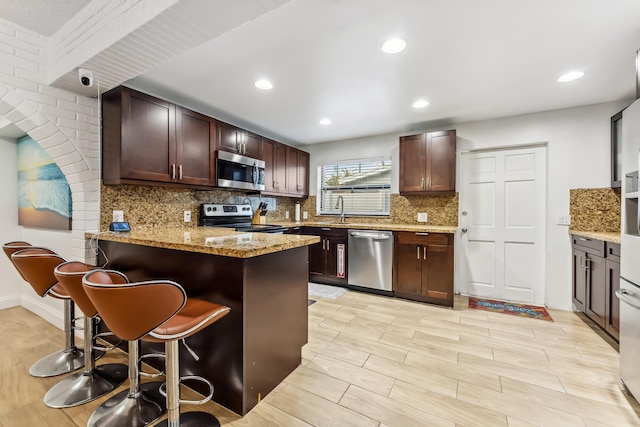 kitchen with kitchen peninsula, a breakfast bar area, appliances with stainless steel finishes, dark brown cabinets, and light stone countertops