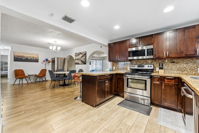 kitchen featuring kitchen peninsula, a breakfast bar area, appliances with stainless steel finishes, light stone countertops, and a chandelier