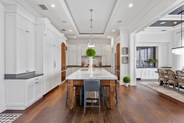 kitchen with crown molding, a kitchen island with sink, hanging light fixtures, white cabinetry, and a raised ceiling