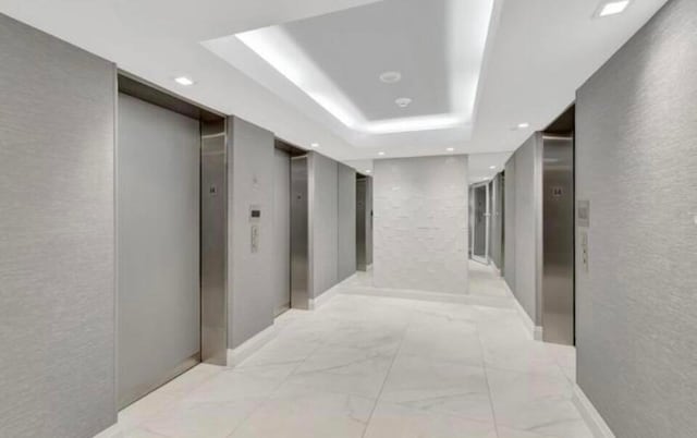 corridor featuring a tray ceiling, elevator, a textured wall, and recessed lighting