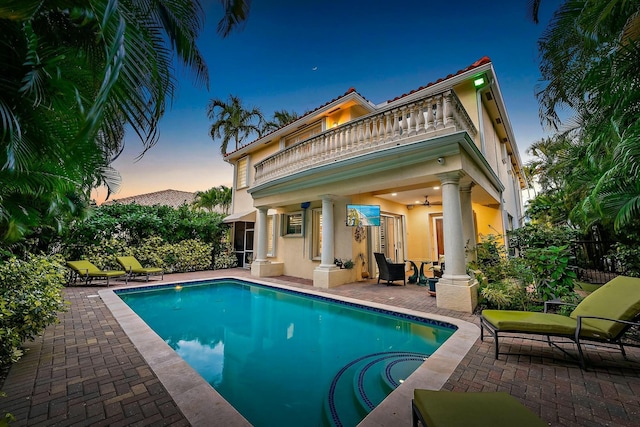 back house at dusk featuring ceiling fan, a patio area, a fenced in pool, and a balcony