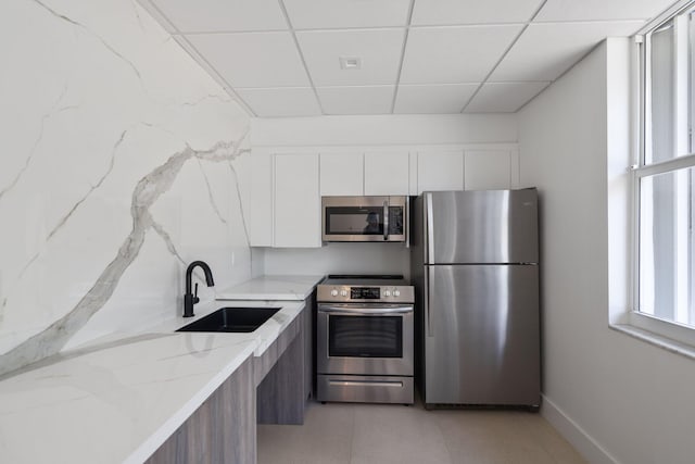 kitchen with appliances with stainless steel finishes, a paneled ceiling, white cabinetry, sink, and light stone counters