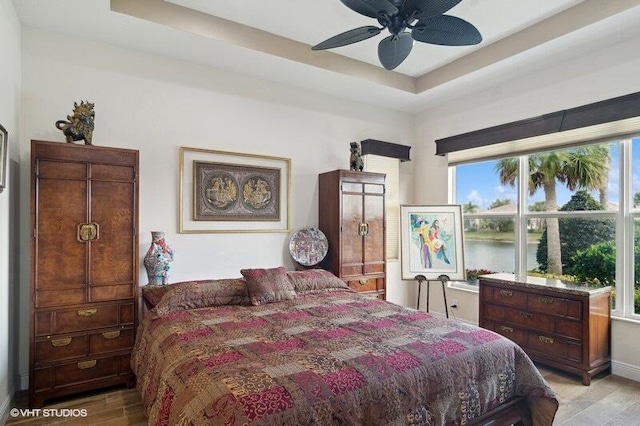 bedroom featuring ceiling fan, light hardwood / wood-style floors, a water view, and a tray ceiling