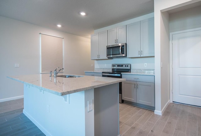 kitchen featuring stainless steel appliances, an island with sink, sink, light stone counters, and gray cabinetry