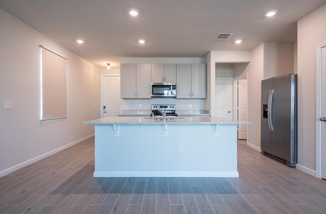 kitchen with gray cabinets, wood-type flooring, a kitchen island with sink, appliances with stainless steel finishes, and light stone counters